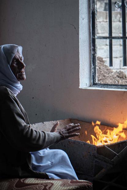 Photograph of a woman in a hijab by a window, a small flame on a surface, another figure in shadow.