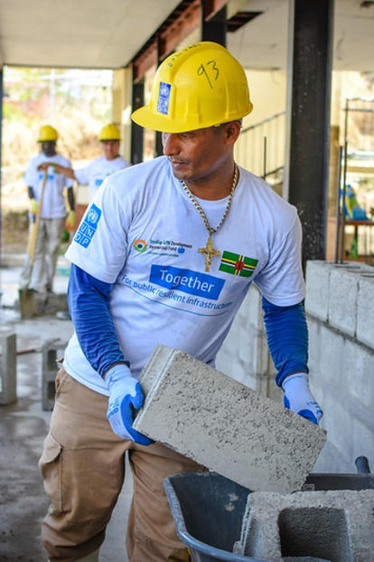 Construction worker in a yellow hard hat holds a concrete block near a wheelbarrow.