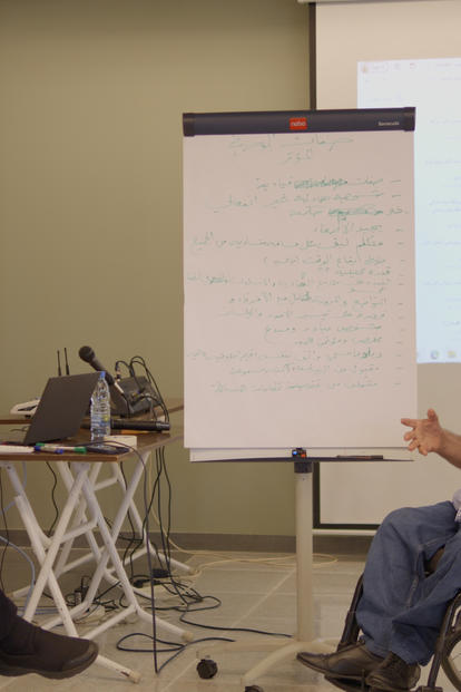 Man in a wheelchair speaking to a small group in a classroom, with projector screen and whiteboard in view.