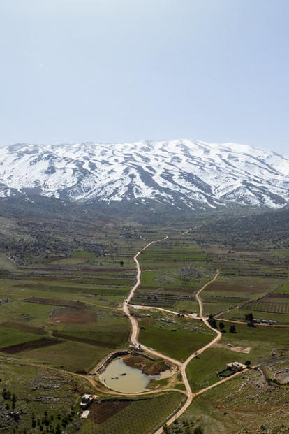 Aerial view of snow-capped mountains beyond rolling green hills with winding dirt roads.