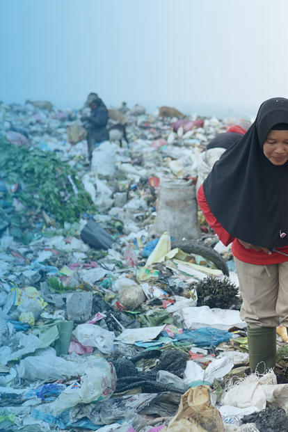 Woman in a red and black hijab scavenges amid a crowded garbage dump.