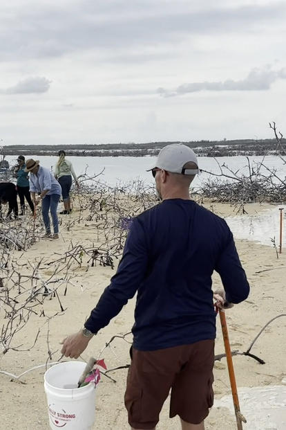 Group of people on a windy beach near dunes; foreground man in blue jacket beside a white bucket.