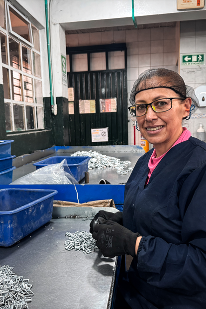 Photograph of a smiling woman in a dark work coat, gloves, and glasses, by blue bins in a workshop.