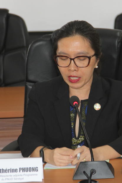 Woman with glasses seated at conference table with microphone, water bottle, and nameplate.
