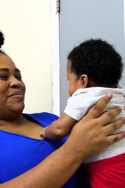 Midwife in blue shirt holds a baby in a clinic with an eye chart on the wall.