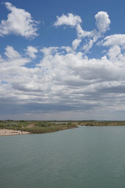 Calm coastal scene with water in foreground, sandy shore and dunes, and a cloudy blue sky.