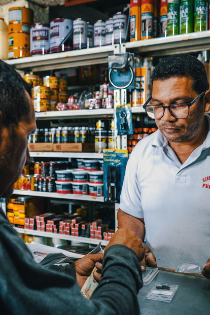Two men at a shop counter; clerk in a white polo assists a customer, shelves stocked behind.