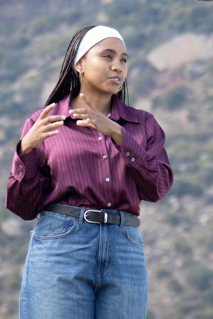 Woman in white headscarf, purple shirt, and jeans poses outdoors with mountains behind.