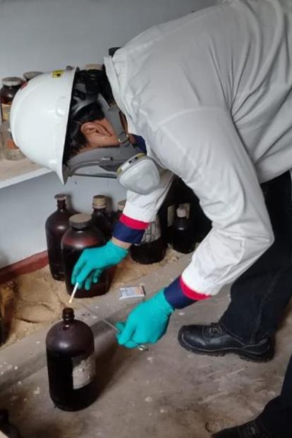 Person in gray sweater bending over a shelf of jars in a kitchen; purple chair nearby.