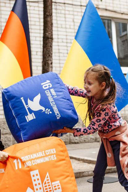 Two kids play with oversized foam blocks outdoors; German, Ukrainian, and UN banners in background.