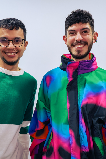 Photograph of two men posing in colorful jackets, smiling; yellow caution sign in background.
