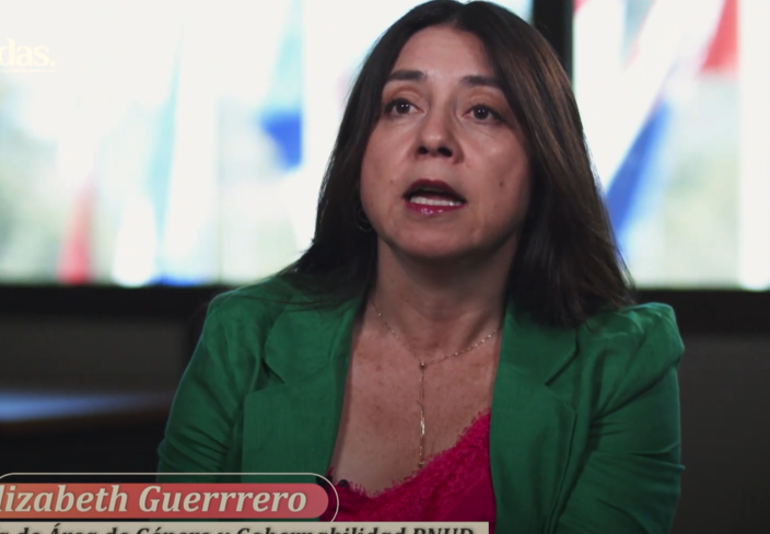 A woman with long dark hair, wearing a green blazer, speaks in front of a window with flags.