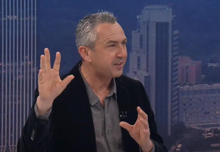 A man with short gray hair gestures while speaking in a studio with a city skyline backdrop.