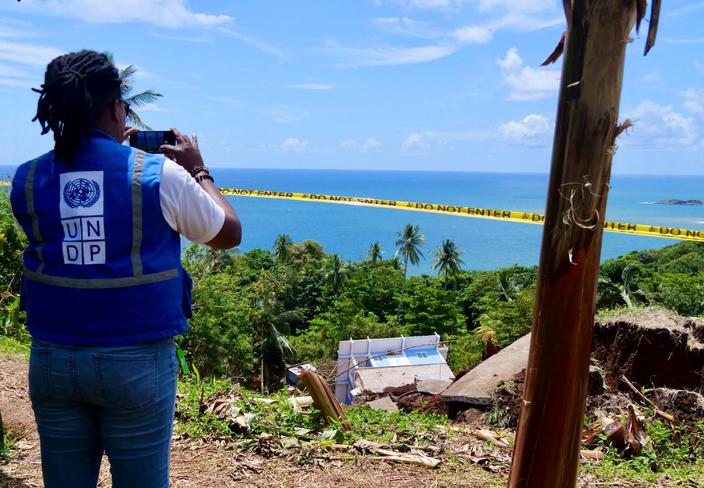 Back view of a person with in a blue UNDP-branded vest, taking a photo of a collapsed house.