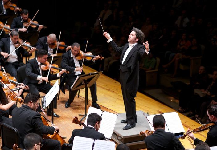 Photograph: conductor raises baton while leading a full orchestra on a lit stage.