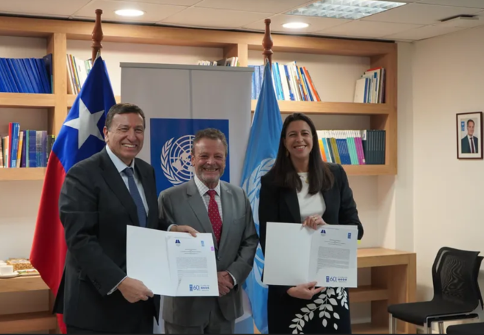 Four adults pose in an office; one holds a certificate, flags and bookshelves in the background.