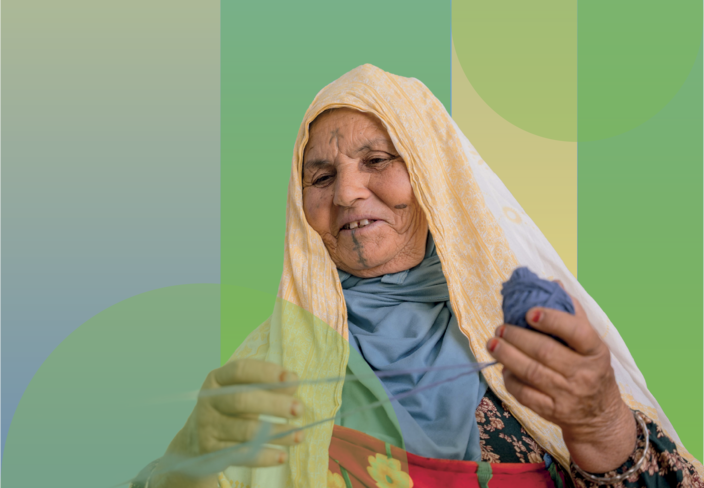 Elderly woman in a beige headscarf and colorful clothes, smiling, holding a glass.