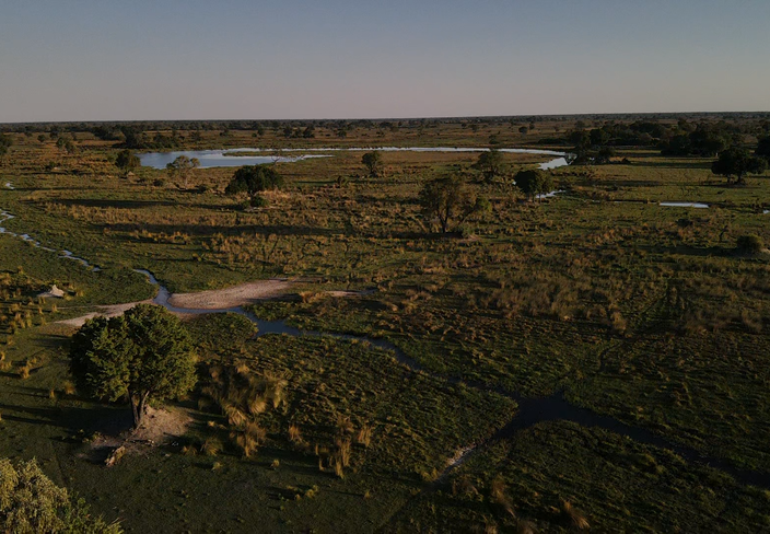 Open rural landscape with green fields, shrubs, scattered houses, and a distant lake.