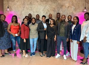 A diverse group of 18 people posing together in a stylish indoor setting with pink lighting.