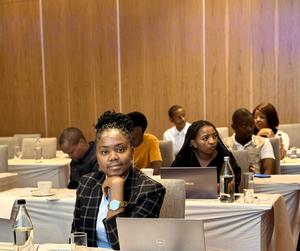 A woman in a blazer sits in a conference room, surrounded by other attendees.