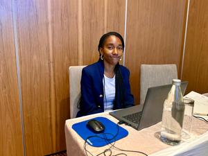 A young woman with long braids smiles while sitting at a desk with a laptop and water bottle.