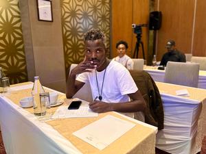 A person with short, curly hair sits at a table in a conference setting, smiling thoughtfully.