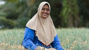 Person in a beige hijab and blue clothing sits in a green crop field with trees in the background.