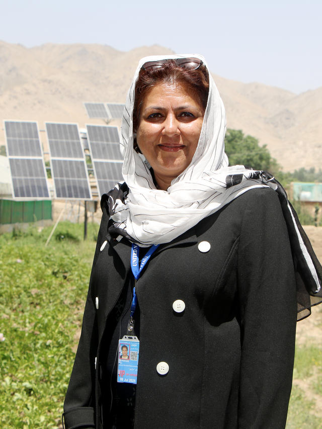 Woman in black abaya with white headscarf and blue lanyard badge, outdoors near solar panels.