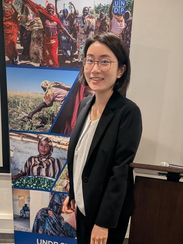 Photograph of a person in a black blazer standing beside a UNDP banner with collage photos.