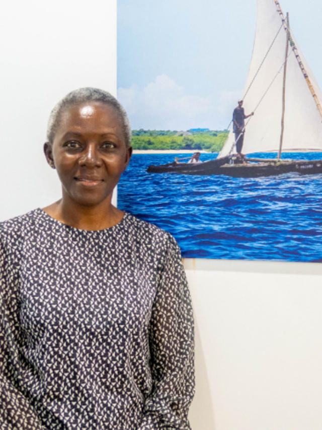Photo of a person in a patterned blouse beside a sailboat painting on a white wall.