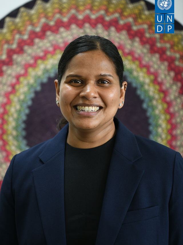 Smiling woman in a navy blazer in front of a colorful woven backdrop; UNDP logo in corner.