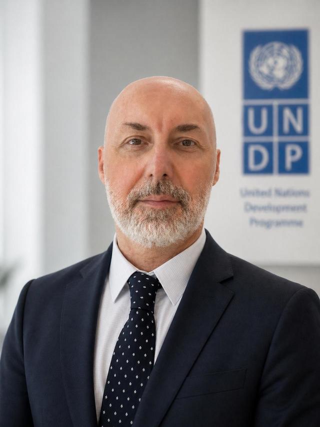 Portrait of a bald, bearded man in a dark suit and navy tie, with UNDP banner.