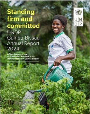 A woman in a green field smiles while watering plants, holding a watering can.