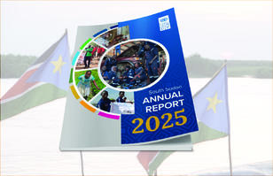 Blue Annual Report 2025 booklet with circular collage on display, flags in background.