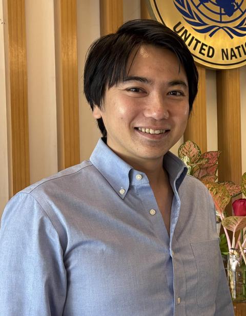 Man with dark hair in a light blue shirt stands indoors with a United Nations emblem behind him.