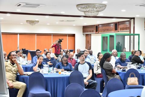 Crowded conference room with attendees at round tables covered in blue cloths; photographer in back.