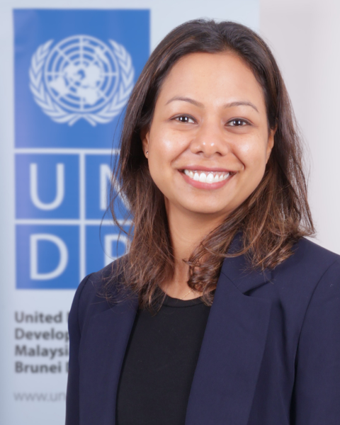 Smiling woman in a navy blazer and black top stands in front of a UNDP banner.