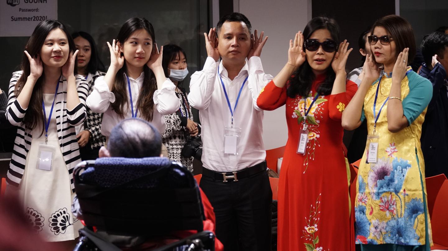 A group of six people engaged in an activity involving hand gestures near their ears during what appears to be a training session. Among them, two women are wearing black glasses, and a man in a wheelchair is facing the group. They are dressed in a mix of traditional Asian garments and casual wear. Photo credits: UNDP Vietnam. 