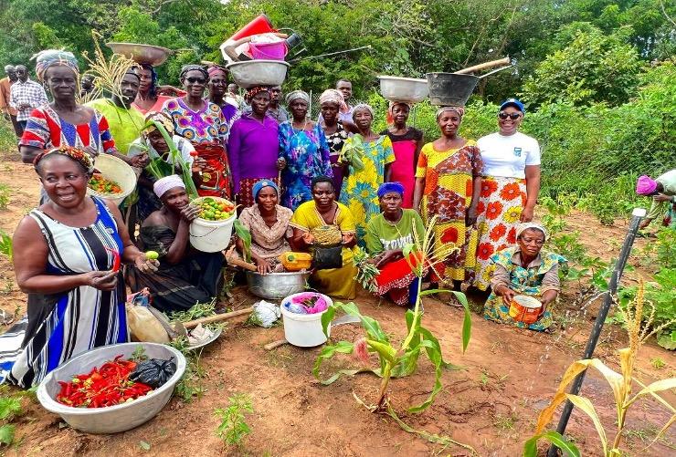A vibrant gathering of women, including women with disabilities in traditional African attire poses with an assortment of fresh produce and cooking utensils. They appear to be in a garden or farm setting, celebrating or participating in a communal activity related to food preparation or harvest. Photo credits: Praise Nutakor, UNDP Ghana. 