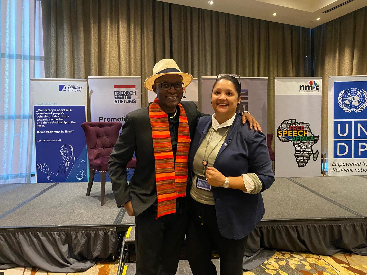 Two individuals pose together smiling at an event, with a stage and banners in the background.