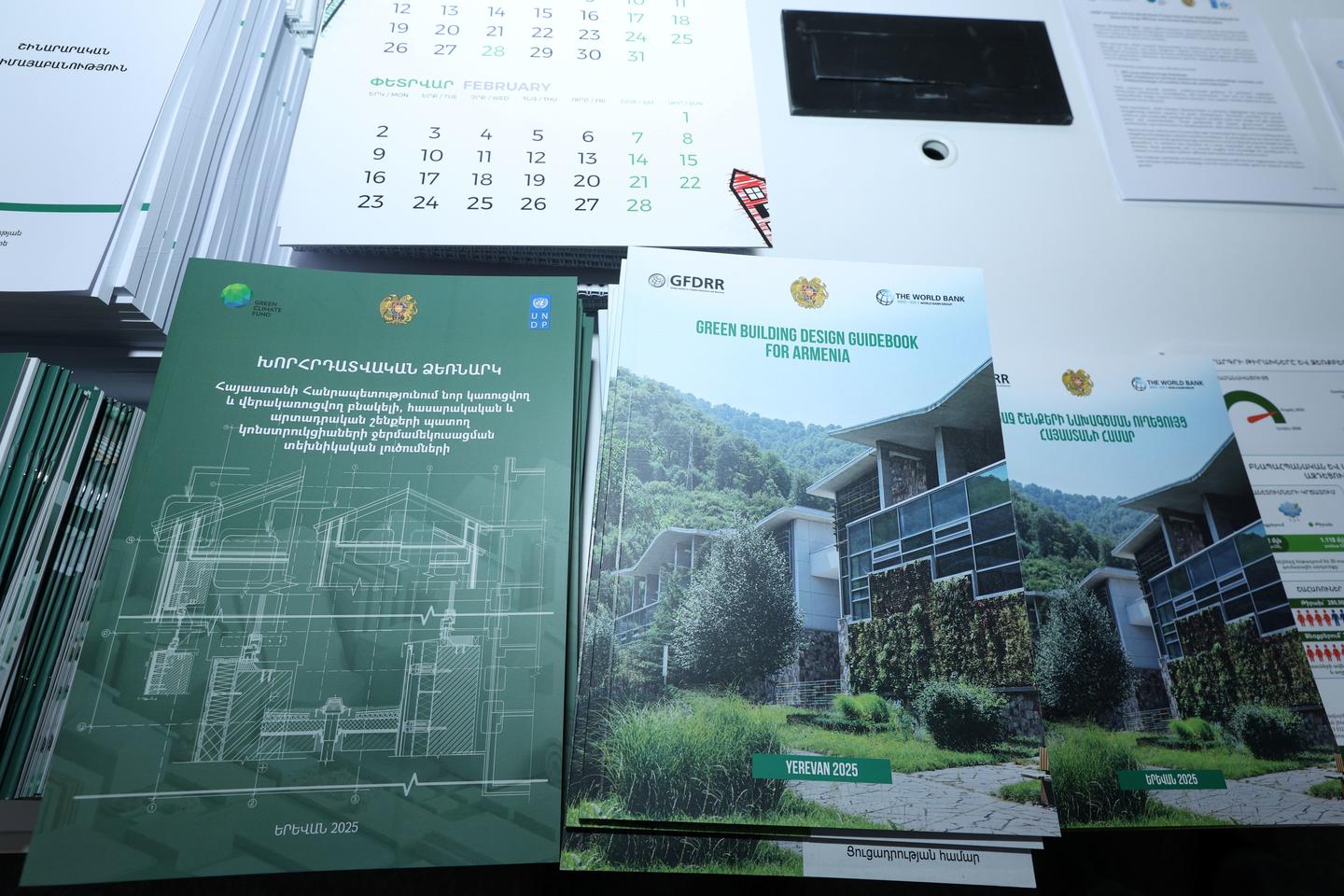 Three green architectural brochures on a desk with a blueprint page and calendar.