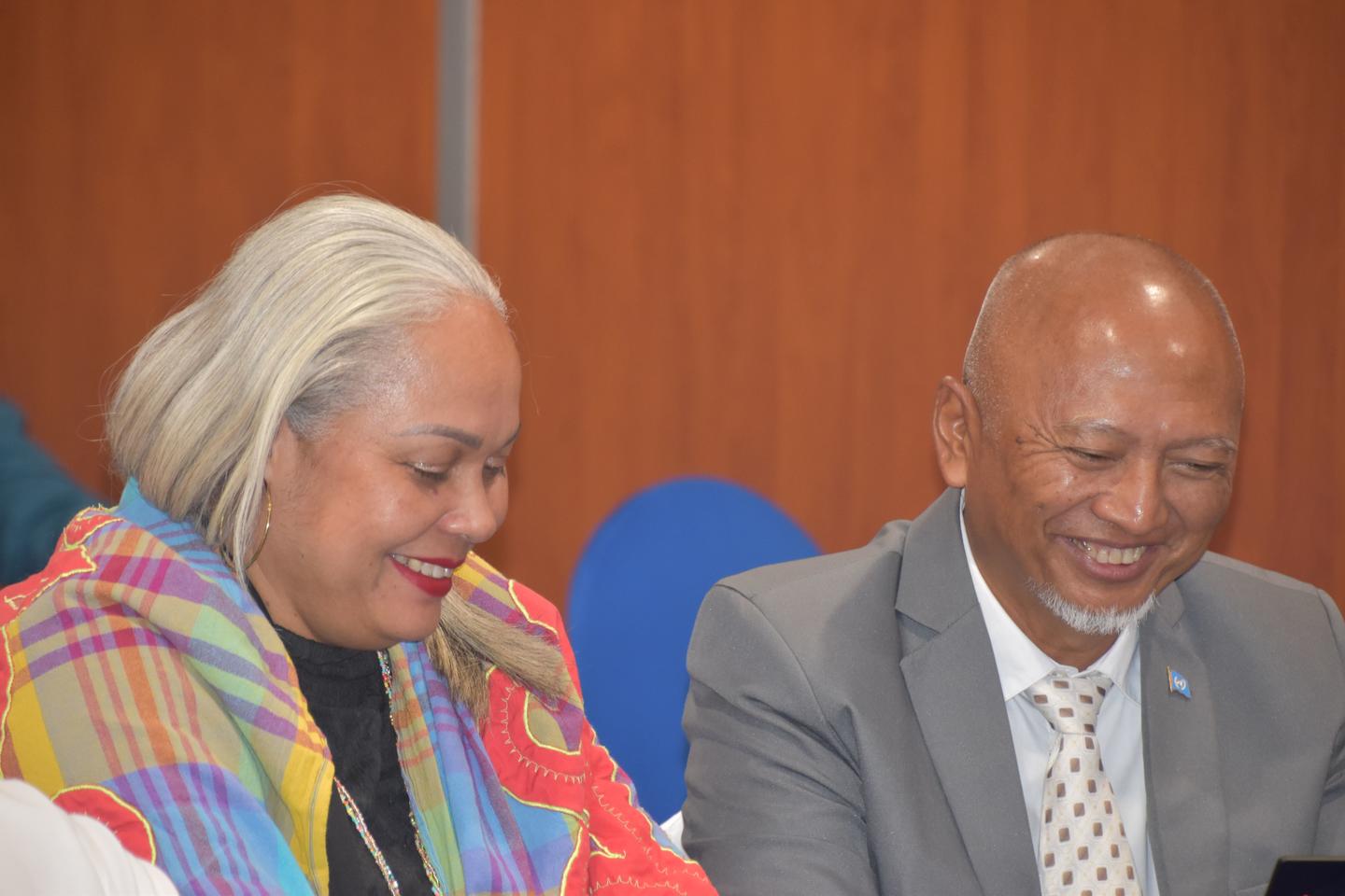 Two people sit at a table in a conference room; left: colorful scarf, right: gray suit.