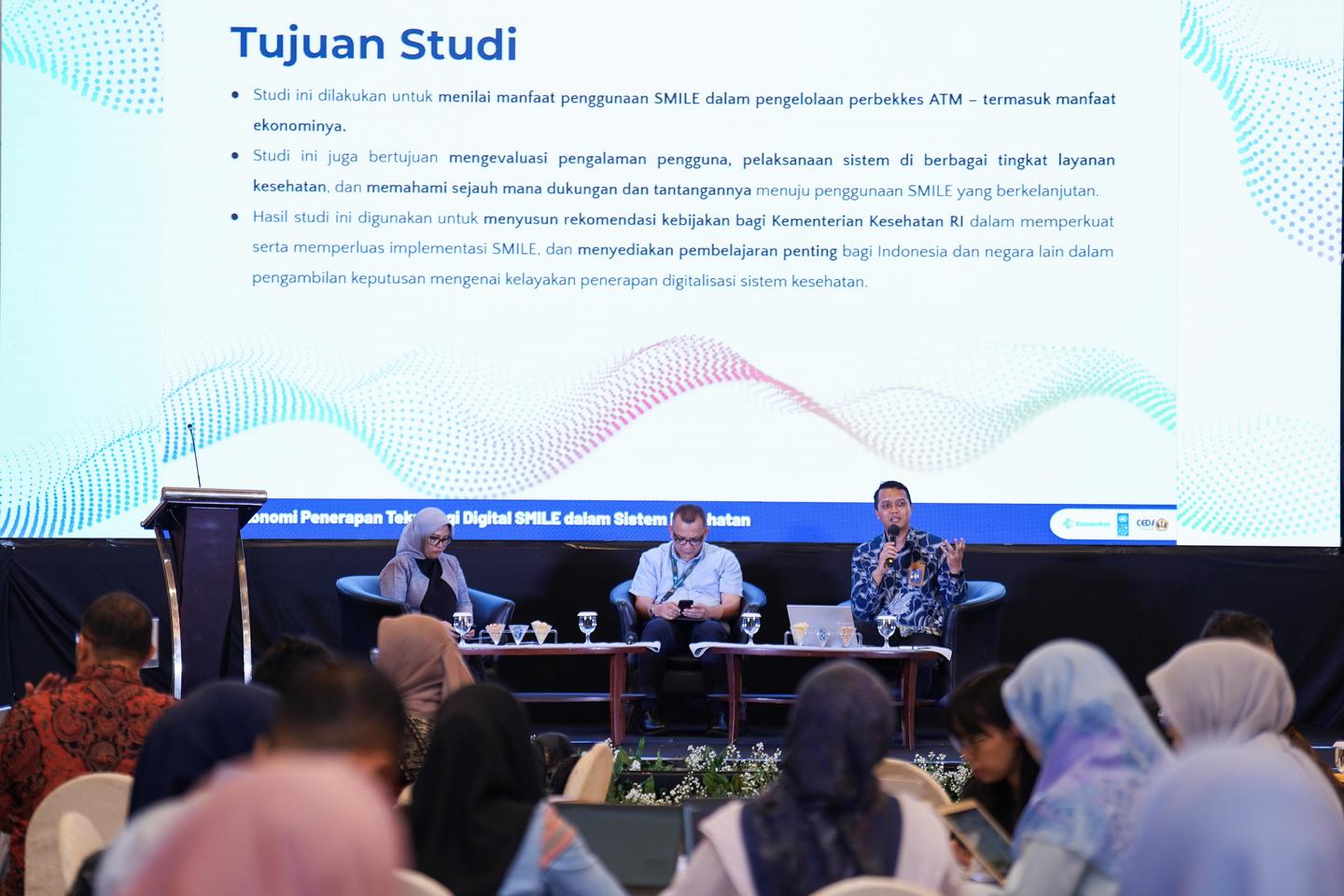 Panel discussion on stage with four speakers, audience listening, slide titled Tujuan Studi in blue.