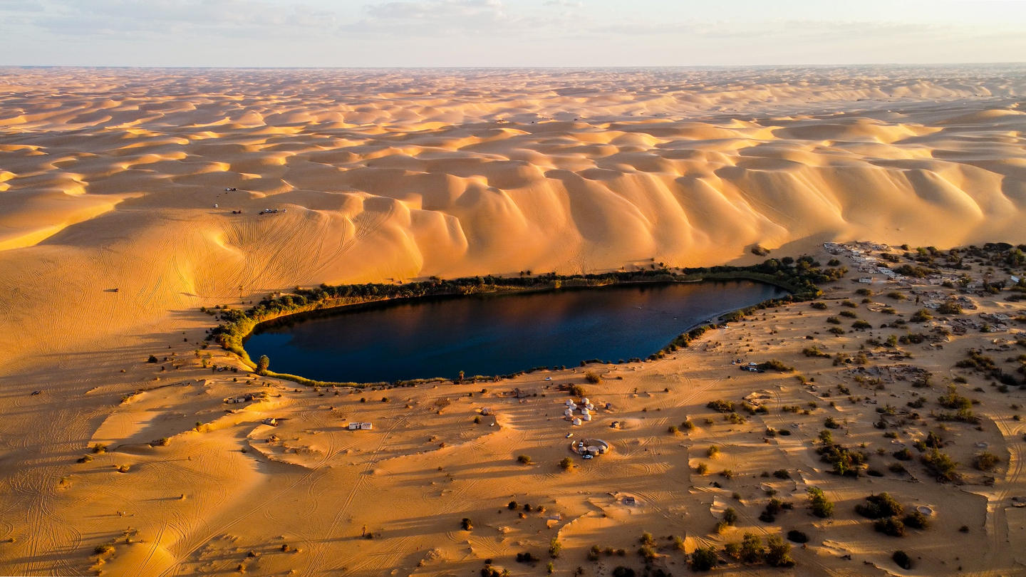 Sunlit desert dunes surround a dark blue crescent-shaped water basin.