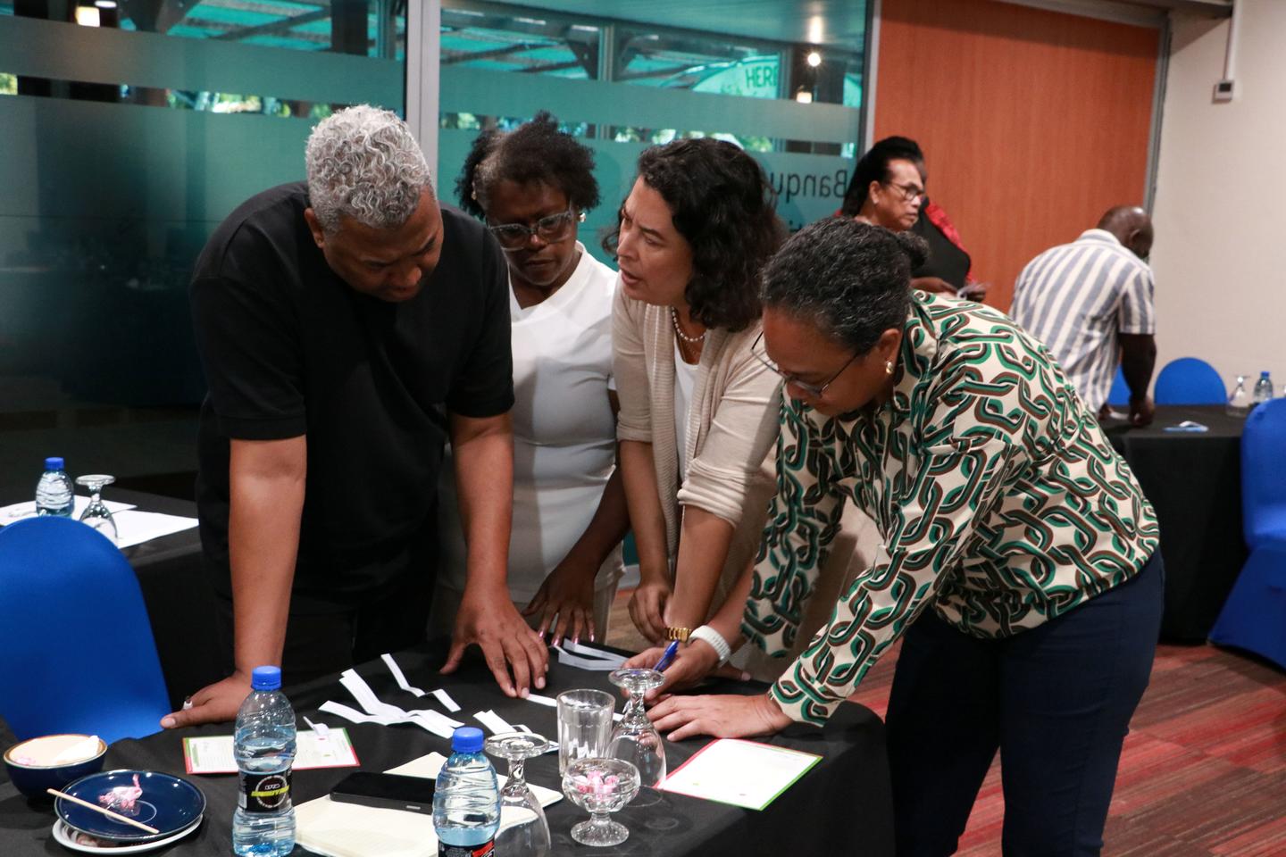 Four people lean over a table of papers and water bottles in a conference room.