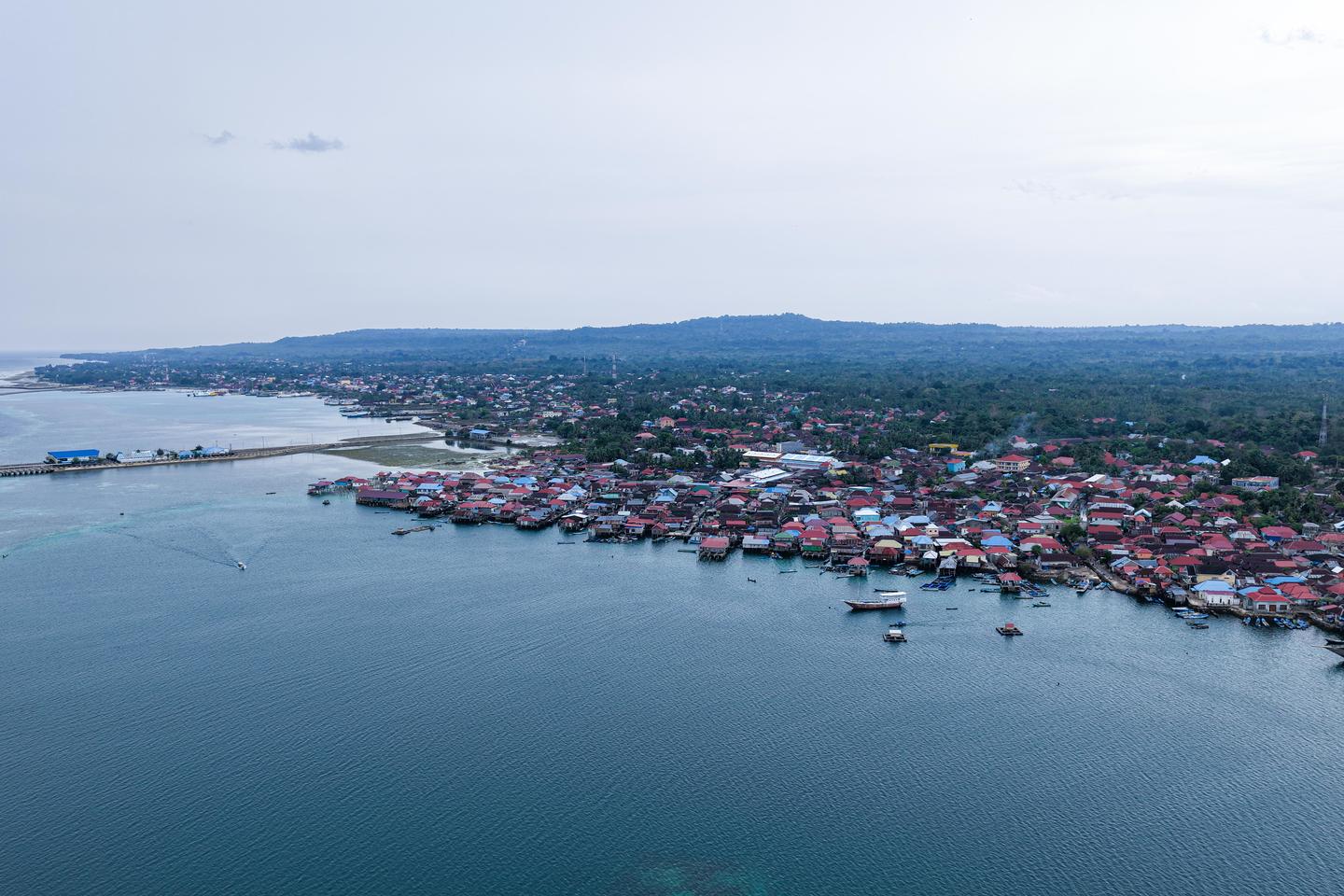 Aerial view of a coastal town with red-roofed houses along a calm bay and harbor boats.