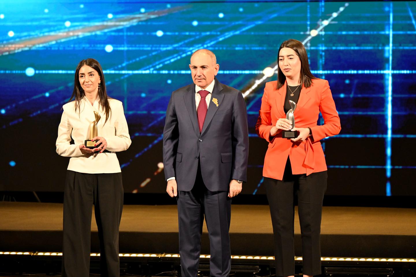 Photograph: three hosts (two women and a man) on stage with a blue digital backdrop.