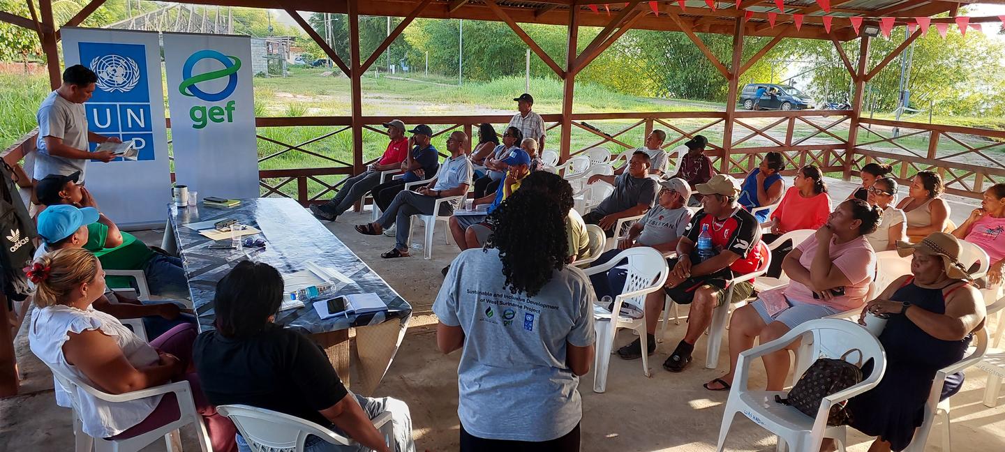 Photograph of an audience seated in a covered outdoor pavilion, listening to speaker with banners.