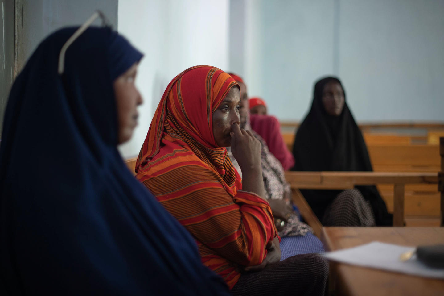 Women in headscarves sit on wooden benches in a classroom; central figure in a red striped scarf.