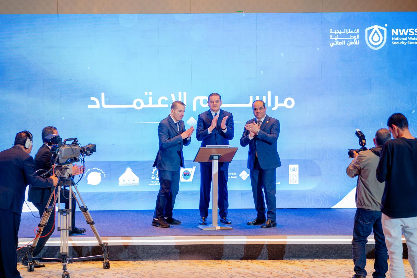Three men in suits on a blue stage shaking hands, with photographers capturing the moment.